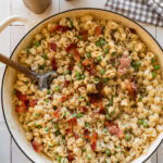 A bowl of peas and bacon pasta with a fork and more ingredients nearby.