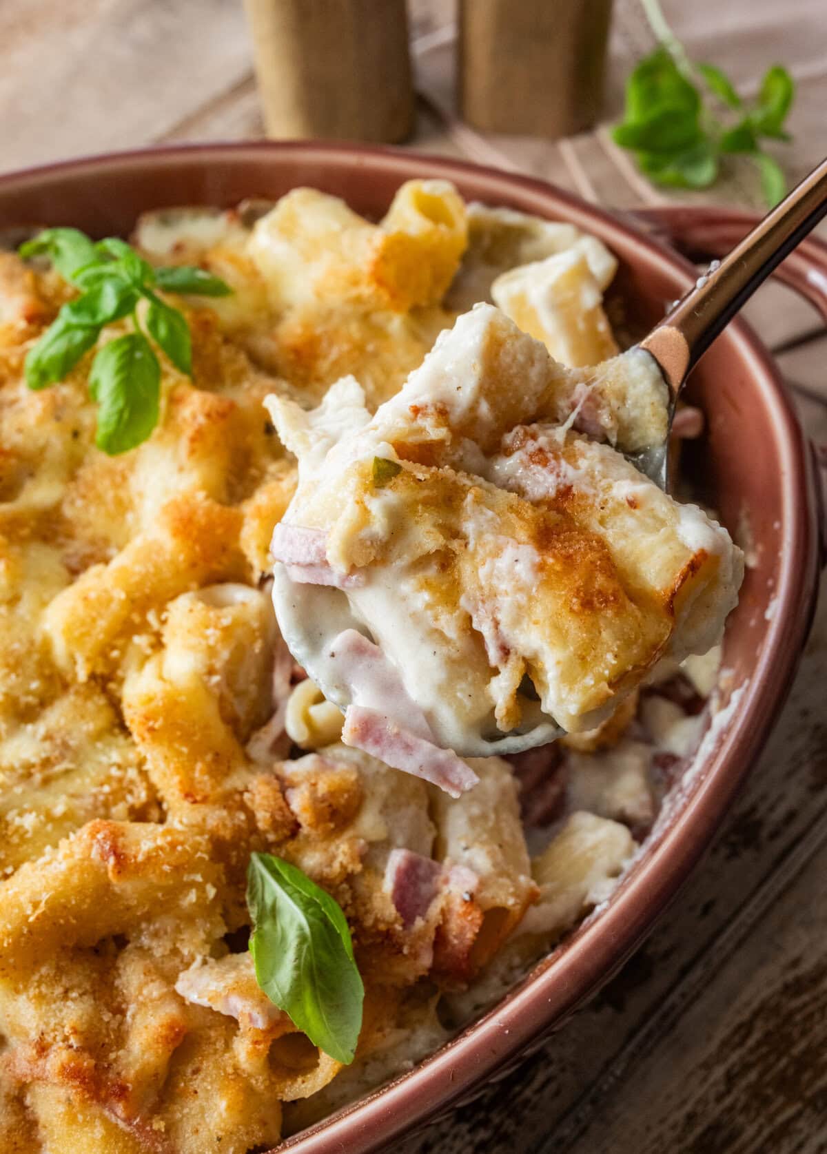 Ham and noodle casserole in a baking dish garnished with basil, and a scoop being taken out.