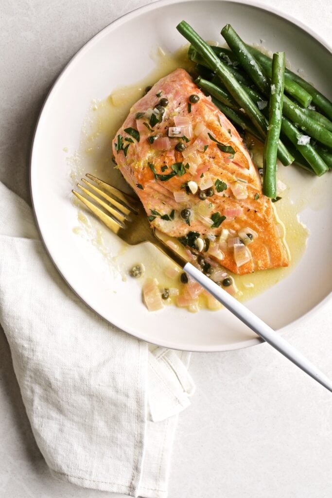 Salmon piccata with sauce and green beans on a plate with a fork.