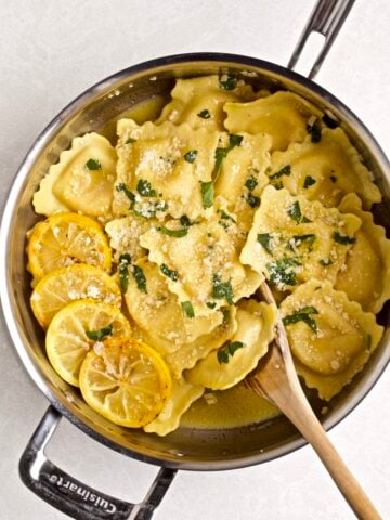 Lobster Ravioli with Lemon Brown Butter Sauce in a pan with a wooden spoon ready to serve. A side of grilled lemons on the side of the pan with the ravioli.