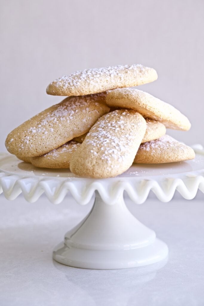 Pile of ladyfinger cookies on a white cake platter.