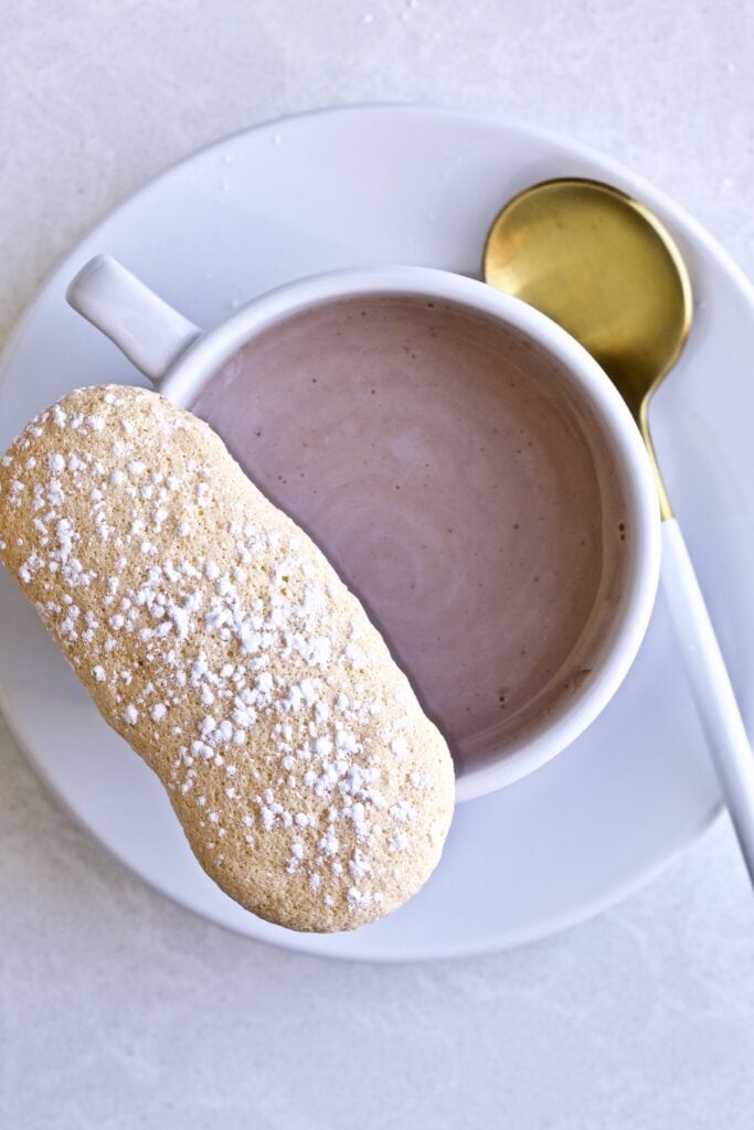 Ladyfinger cookie on the rim of a cup of hot chocolate on a saucer with a spoon.