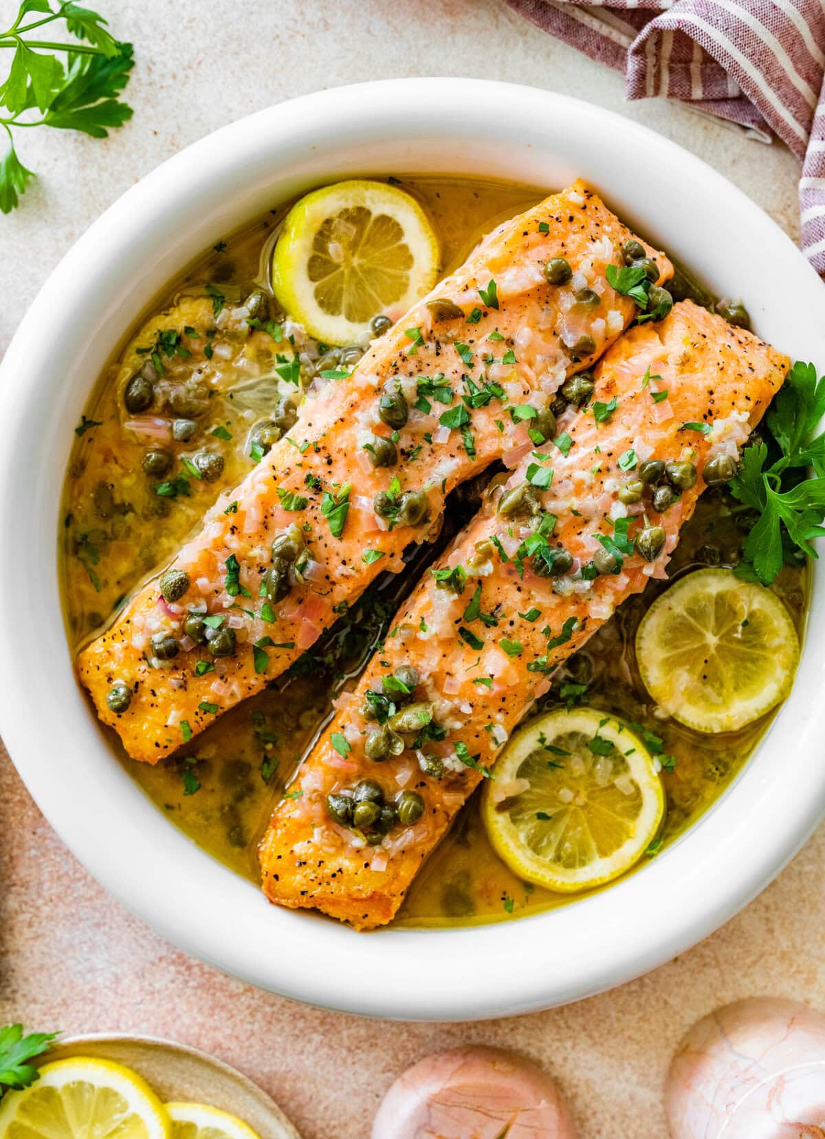 Salmon piccata in a white bowl with slices of lemon and parsley sprinkled on top.