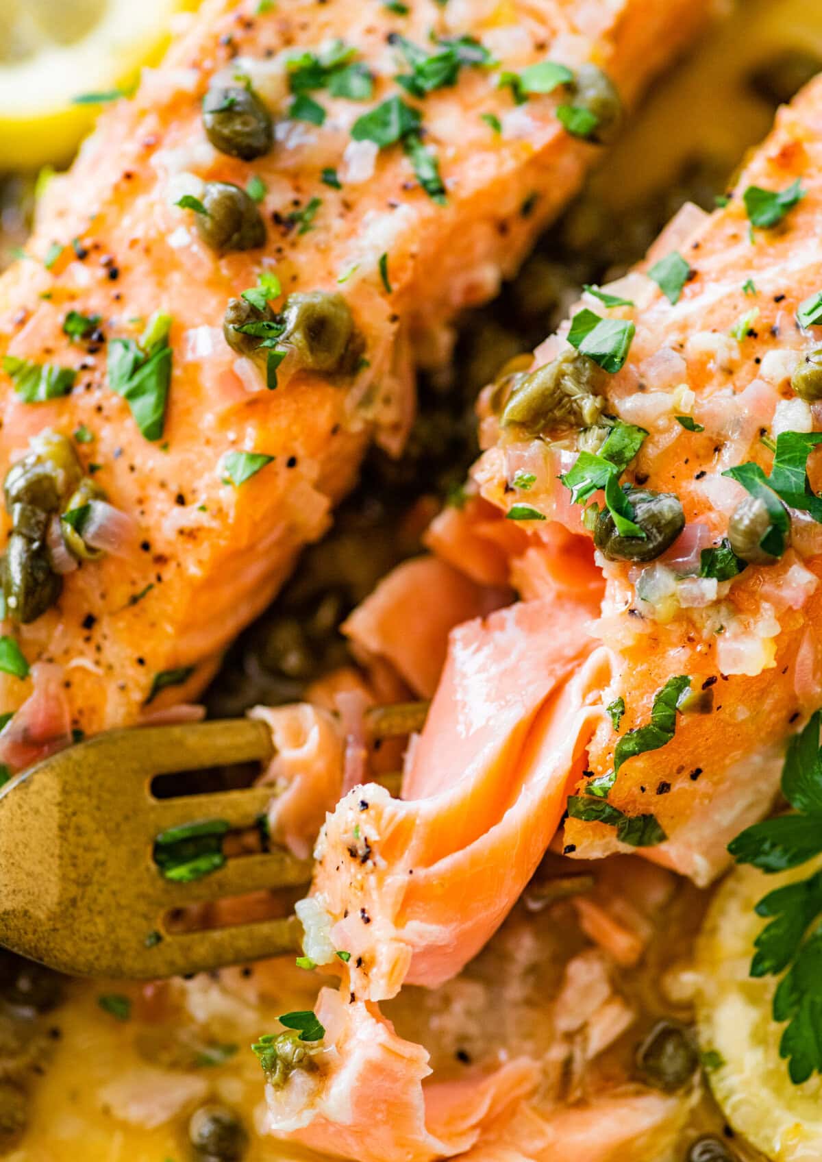 A fork taking a bite from a salmon fillet with caper sauce.