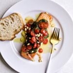 Salmon Pesto Recipe- on a plate with cherry tomatoes and pine nuts on top. A fork and slice of fresh bread on the plate.