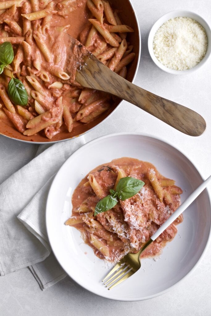 Penne in pink sauce on a plate with a fork in it. The pan of the pasta and sauce is in the background.