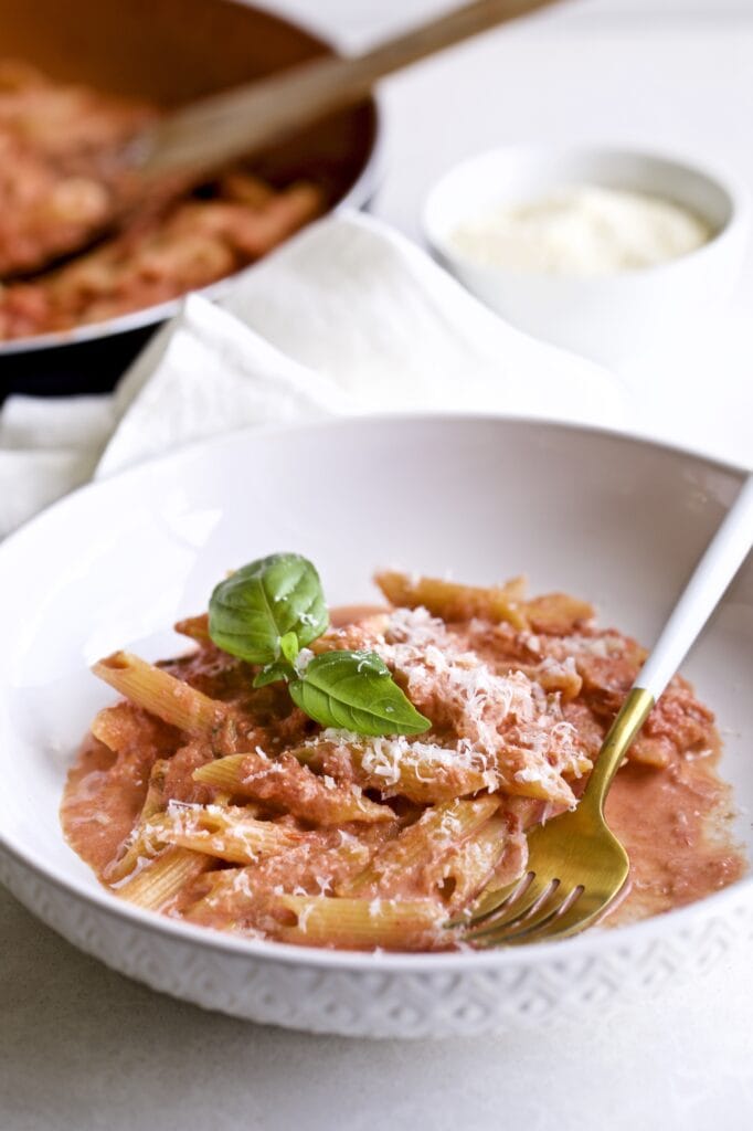 Penne in pink sauce on a plate with a fork in it. The pan of the pasta and sauce is in the background.