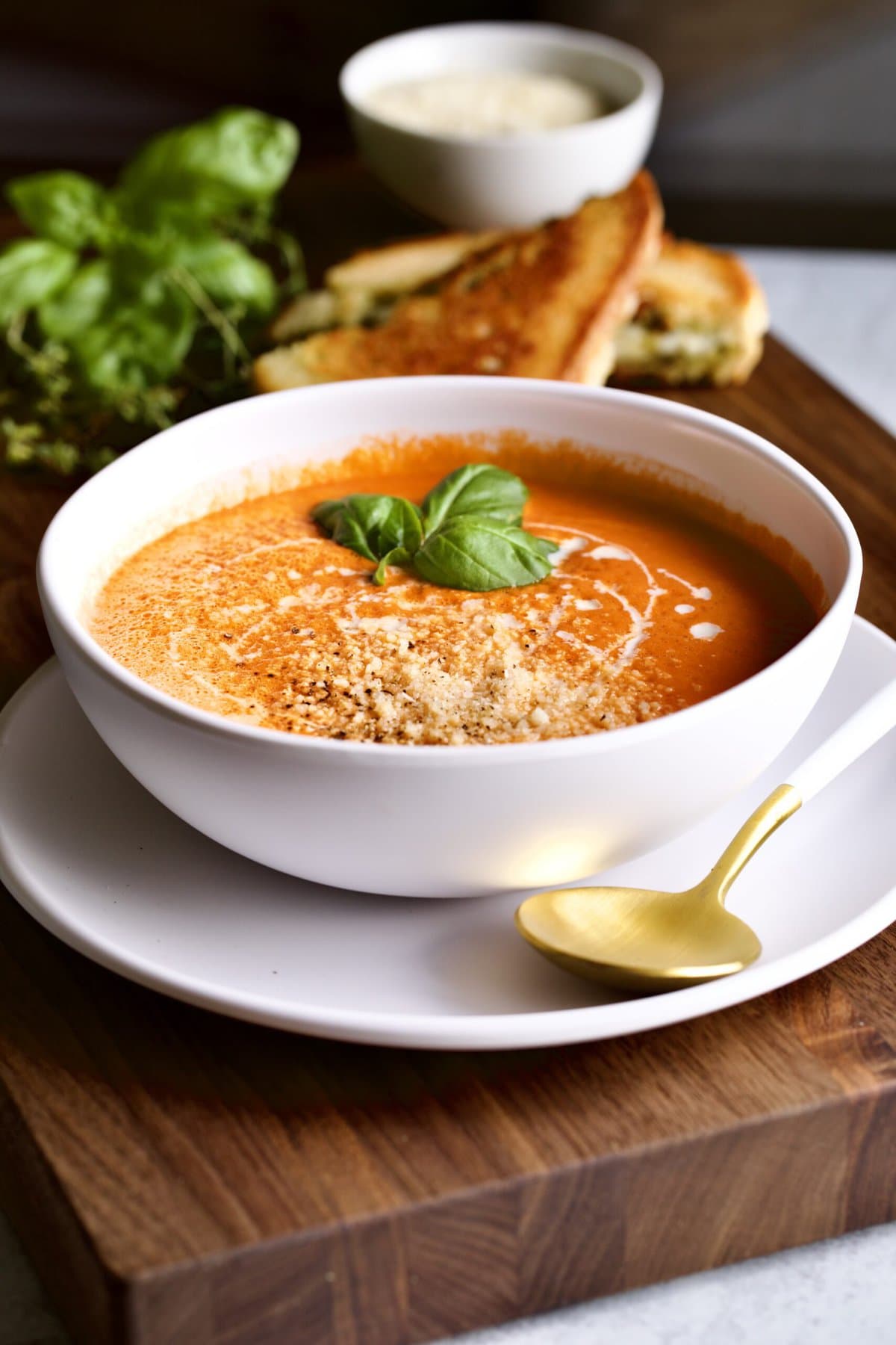 tomato soup in bowl with spoon on a plate. pesto grilled cheese in the background.
