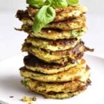 stacks of zucchini fritters on a plate. Basil leaf on top.