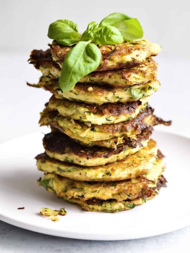stacks of zucchini fritters on a plate. Basil leaf on top.