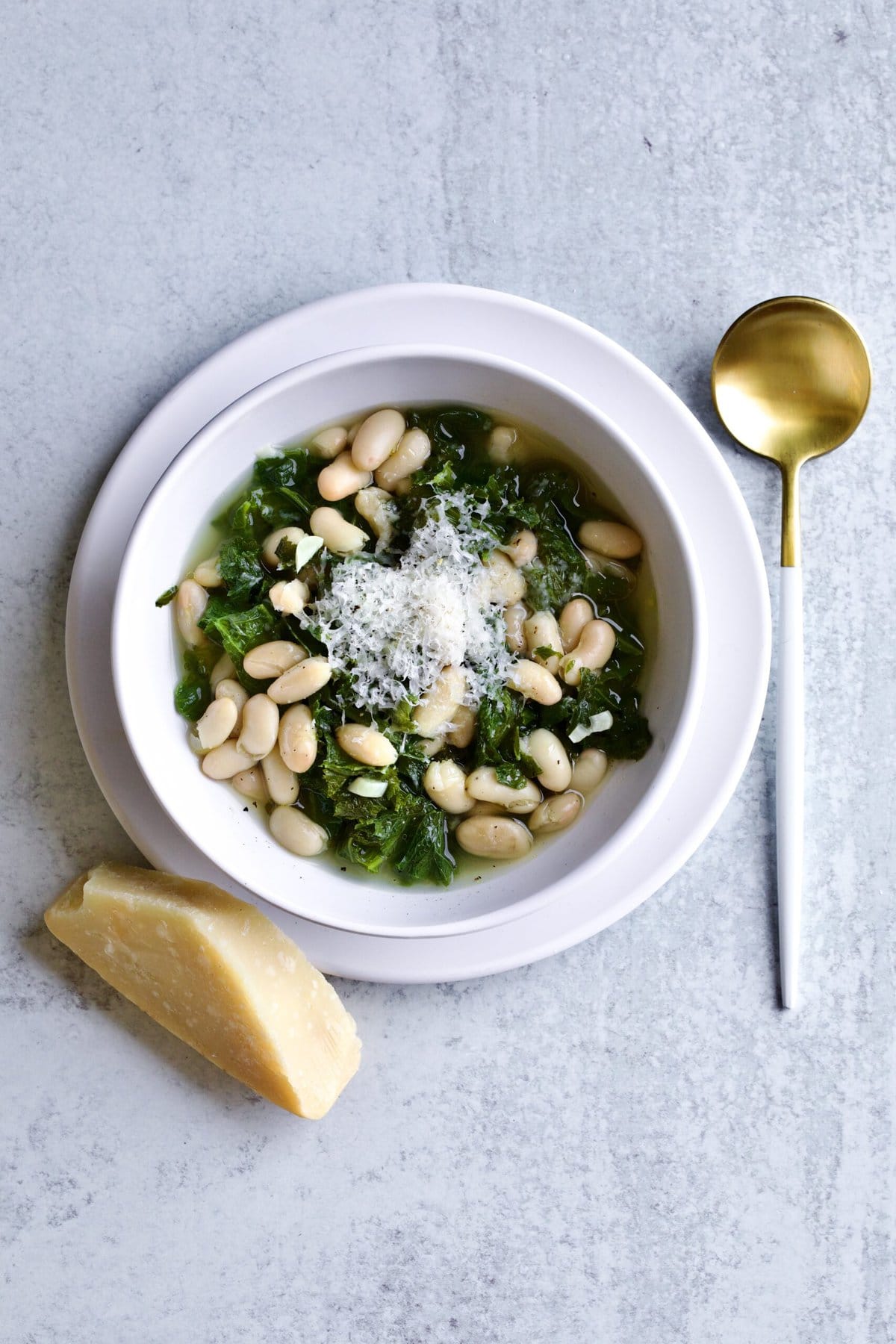 Italian beans and greens soup in a bowl with a gold and white spoon. Piece of parm cheese next to bowl.