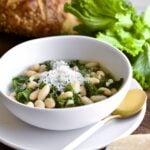 Italian beans and greens soup in a bowl with crusty bread and escarole leaves in background.