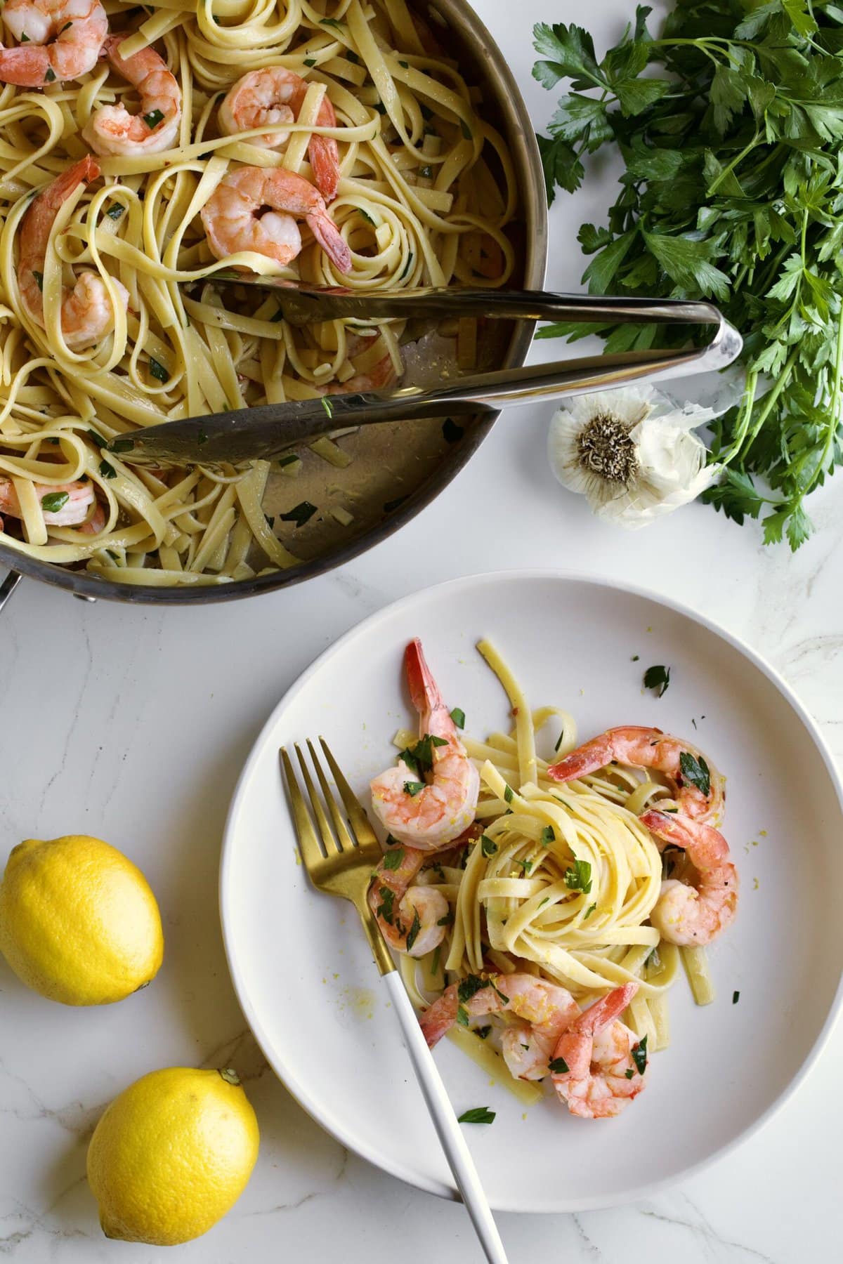 shrimp scampi fettuccine on a plate with parsley and a fork and serving pan in background. Lemon and parsley displayed. 