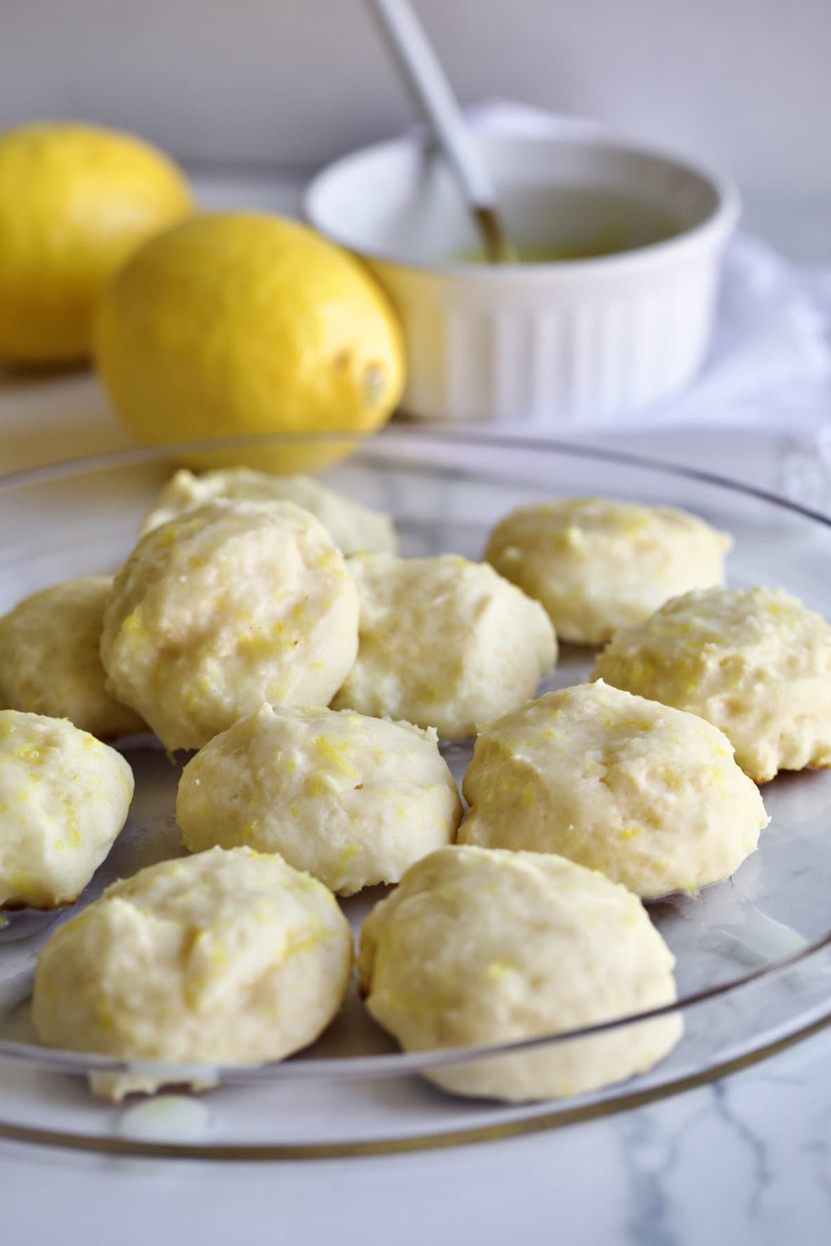 lemon ricotta cookies on a glass plate with lemons in the background.