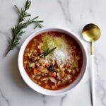 bowl of pasta e fagioli with a spoon and sprig of rosemary on side.