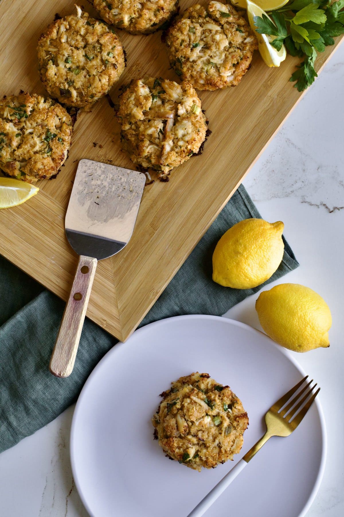 crab cakes displayed on wooden board with lemon and parsley. one crab cake on a white plate.