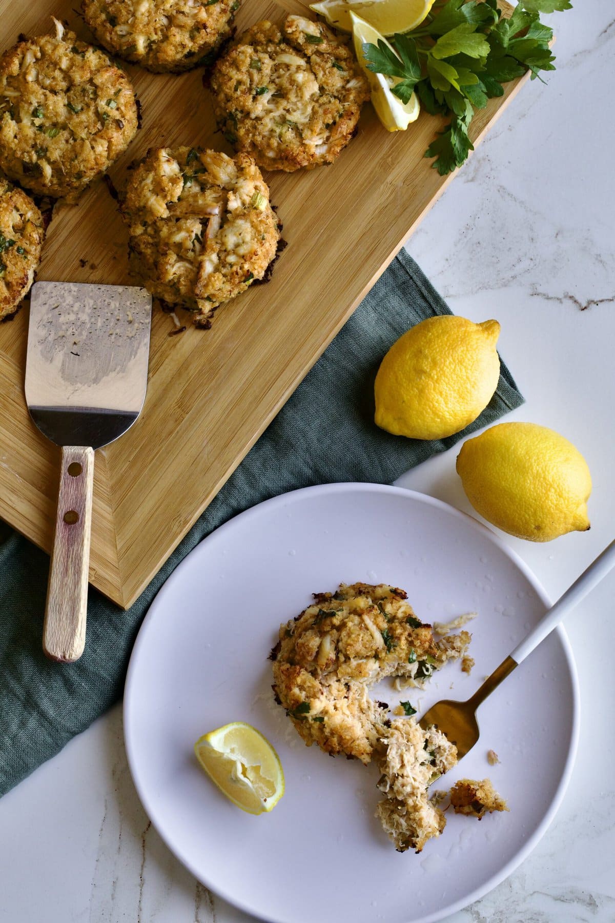 crab cakes displayed on wooden board with lemon and parsley. one crab cake on a white plate. with bite taken out of it.