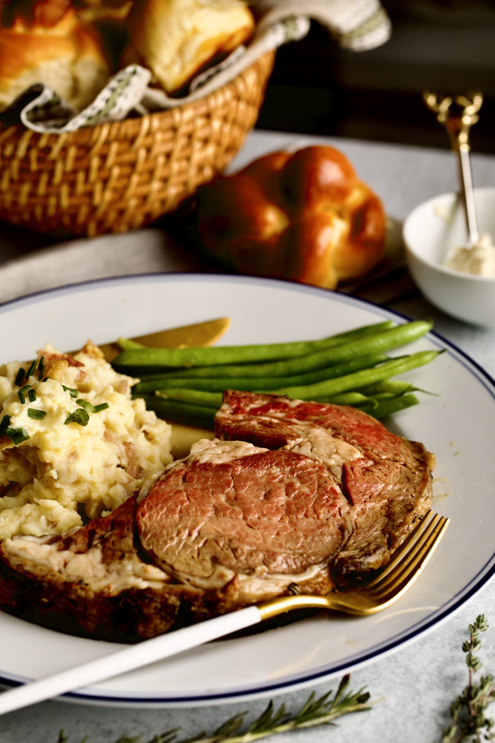 prime rib slice on a plate with garlic red shin mashed potatoes and green beans. Horseradish and dinner roll in background.