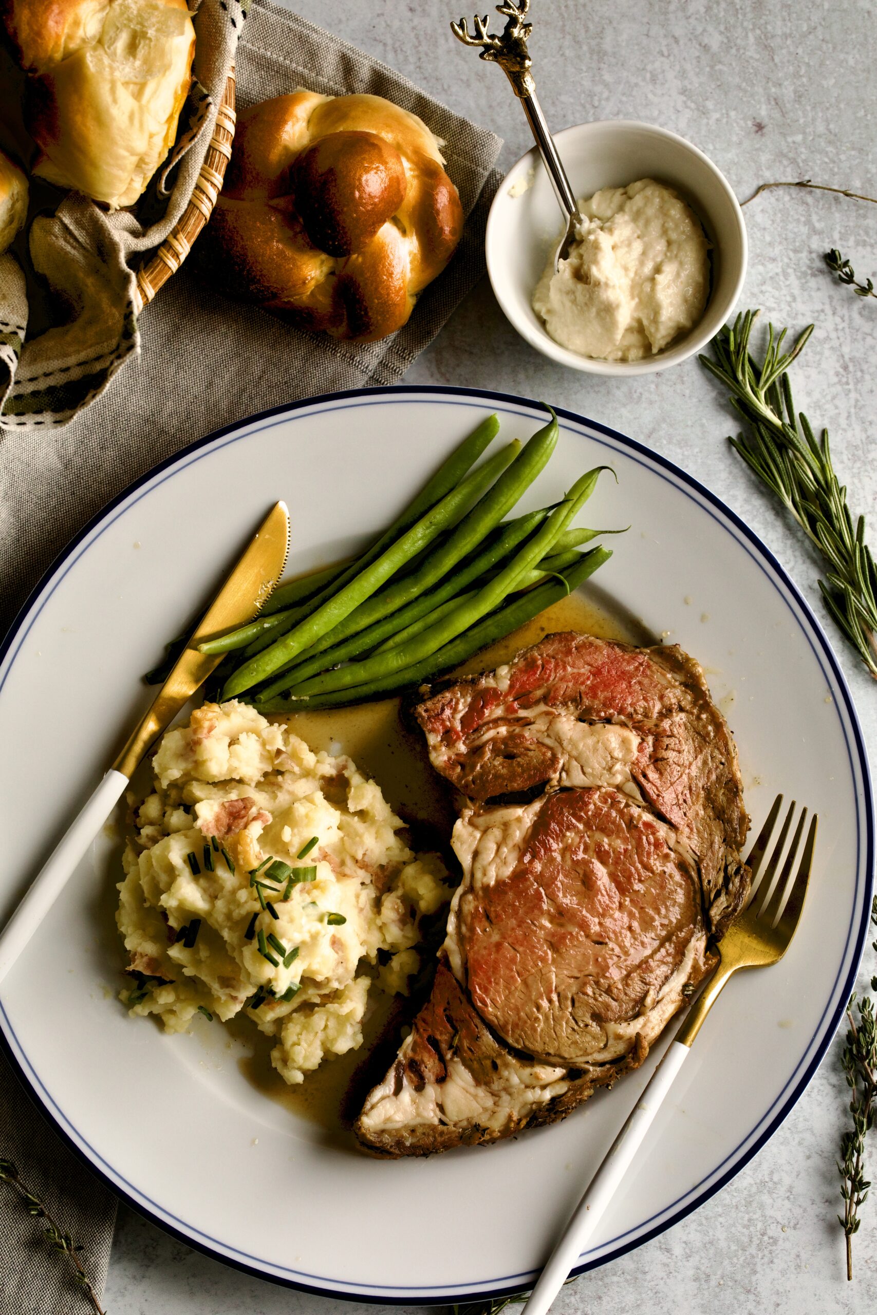 prime rib slice on a plate with garlic red shin mashed potatoes and green beans. Horseradish and dinner roll in background.