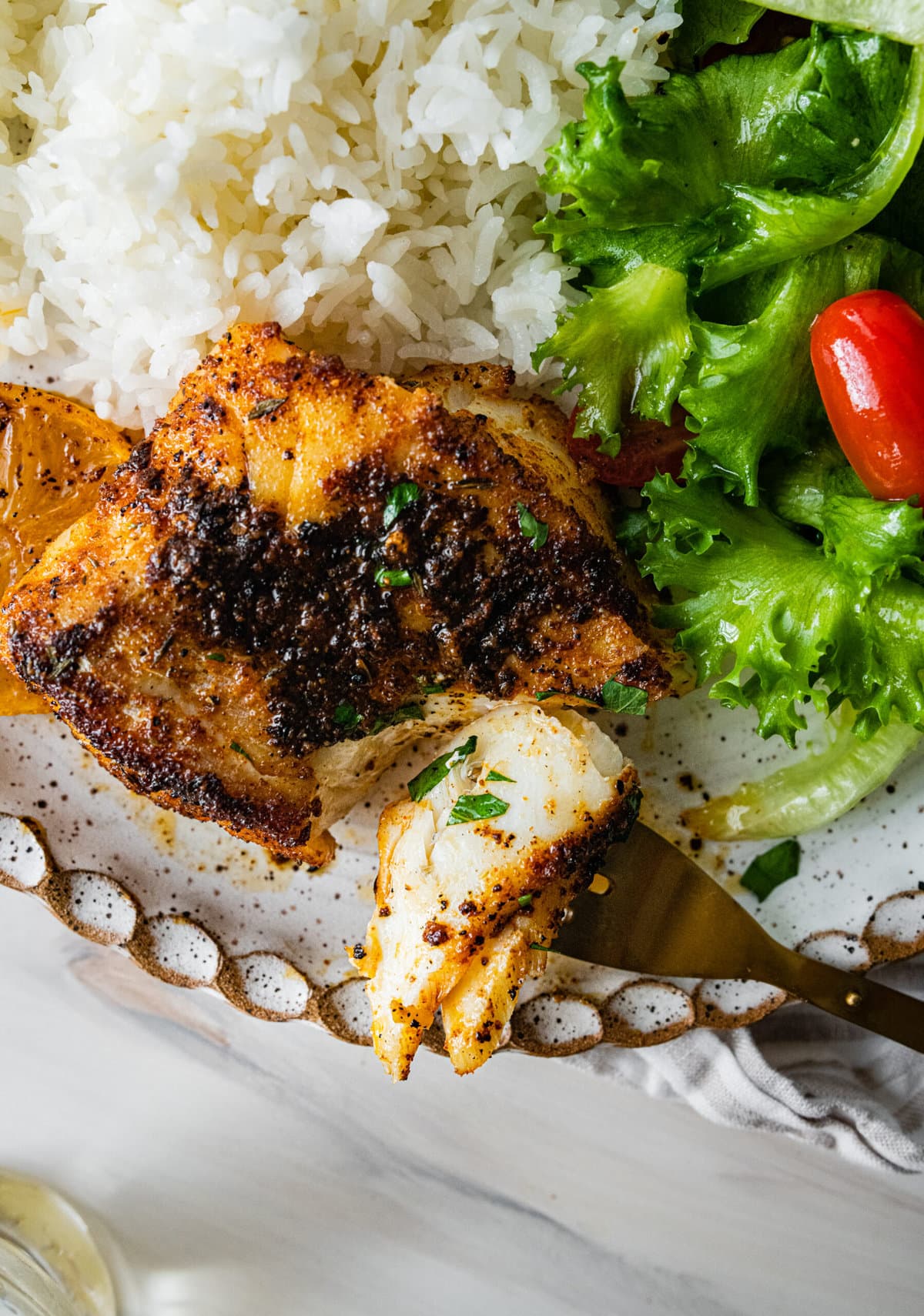 platted blackened cod with white rice and green salad with tomatoes. Bite of flaky fish on fork.
