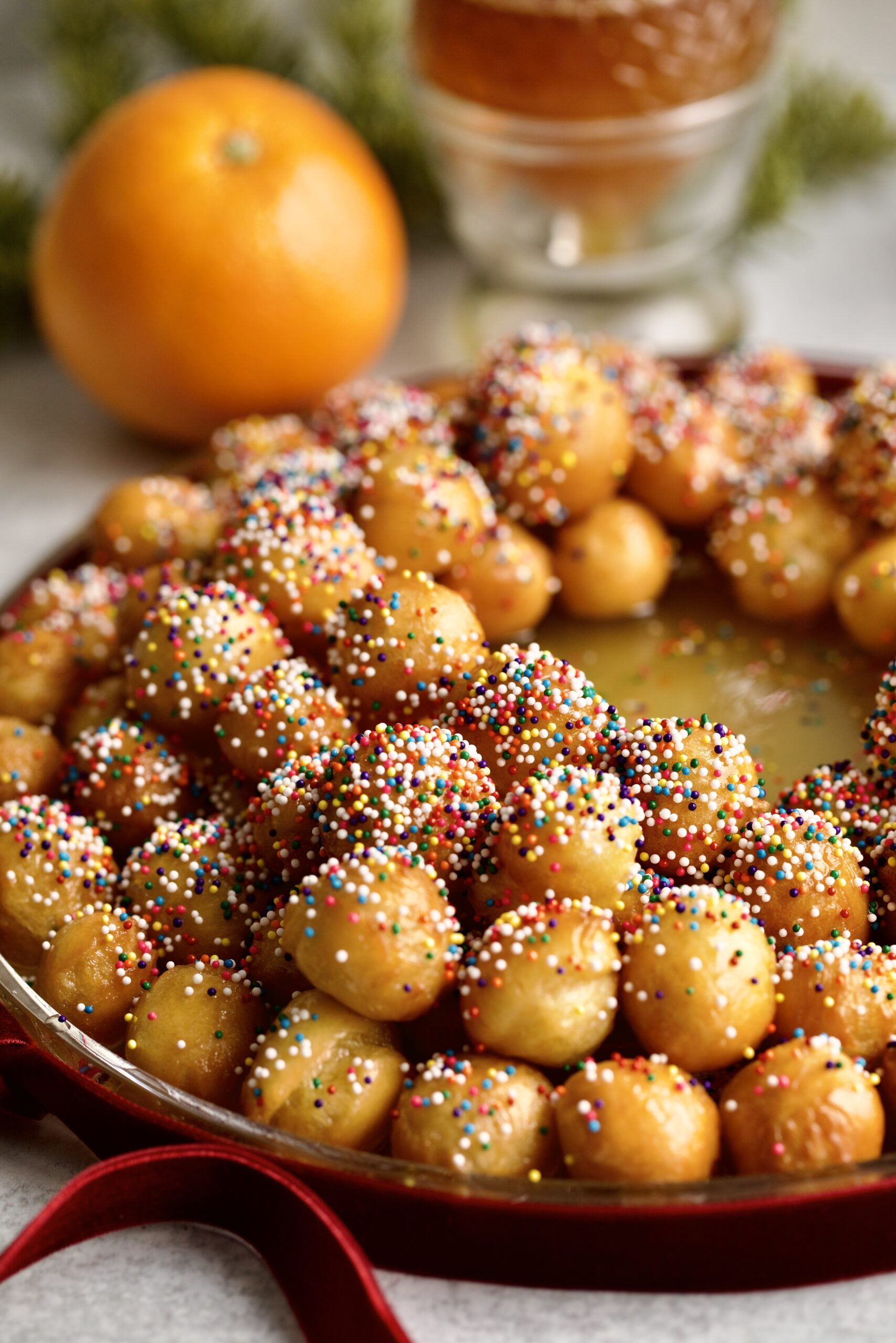 struffoli honey dough balls with sprinkles on a serving platter- wreath shape. Orange and Christams ribbon on background.