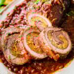 Partially-sliced beef braciole over sauce on a white platter.