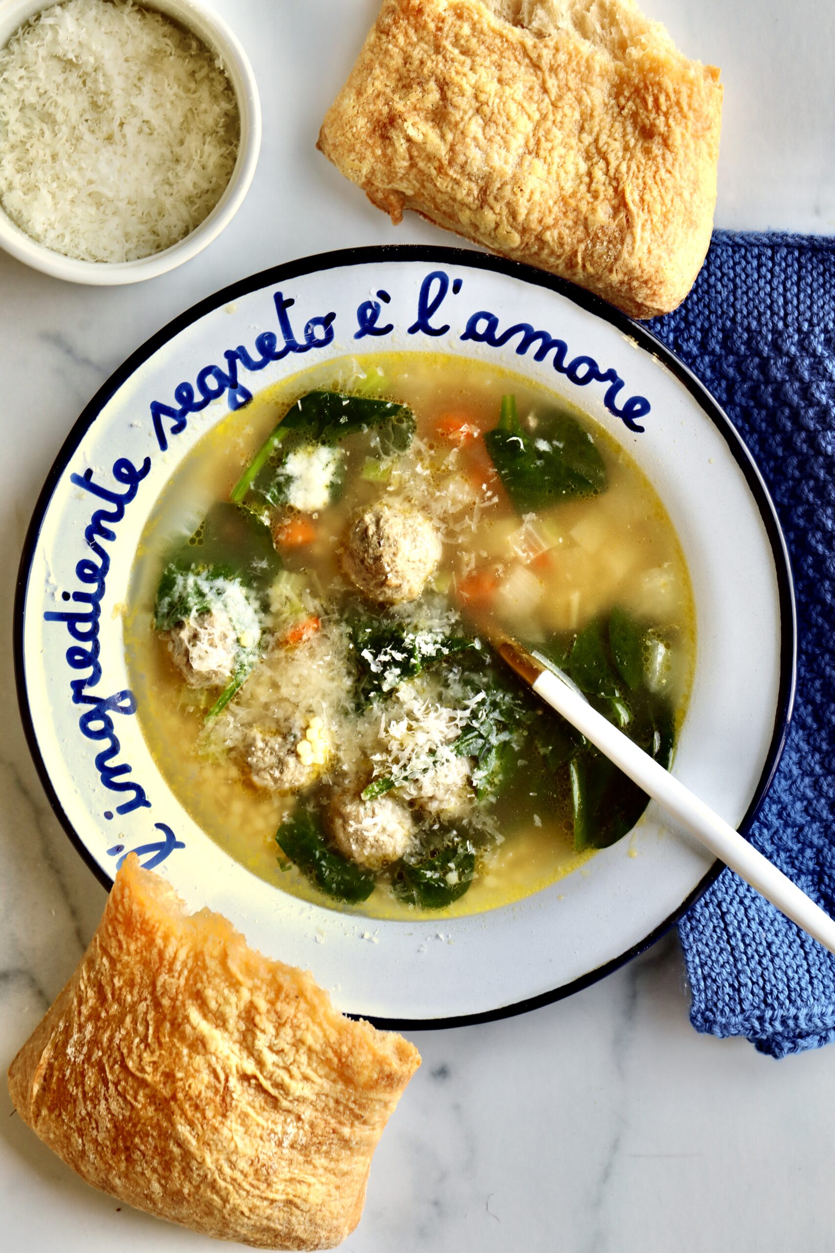 Classic Italian Wedding Soup Recipe with a bowl of freshly grated parmigiano in the background. Blue hand knit napkin next to bowl.