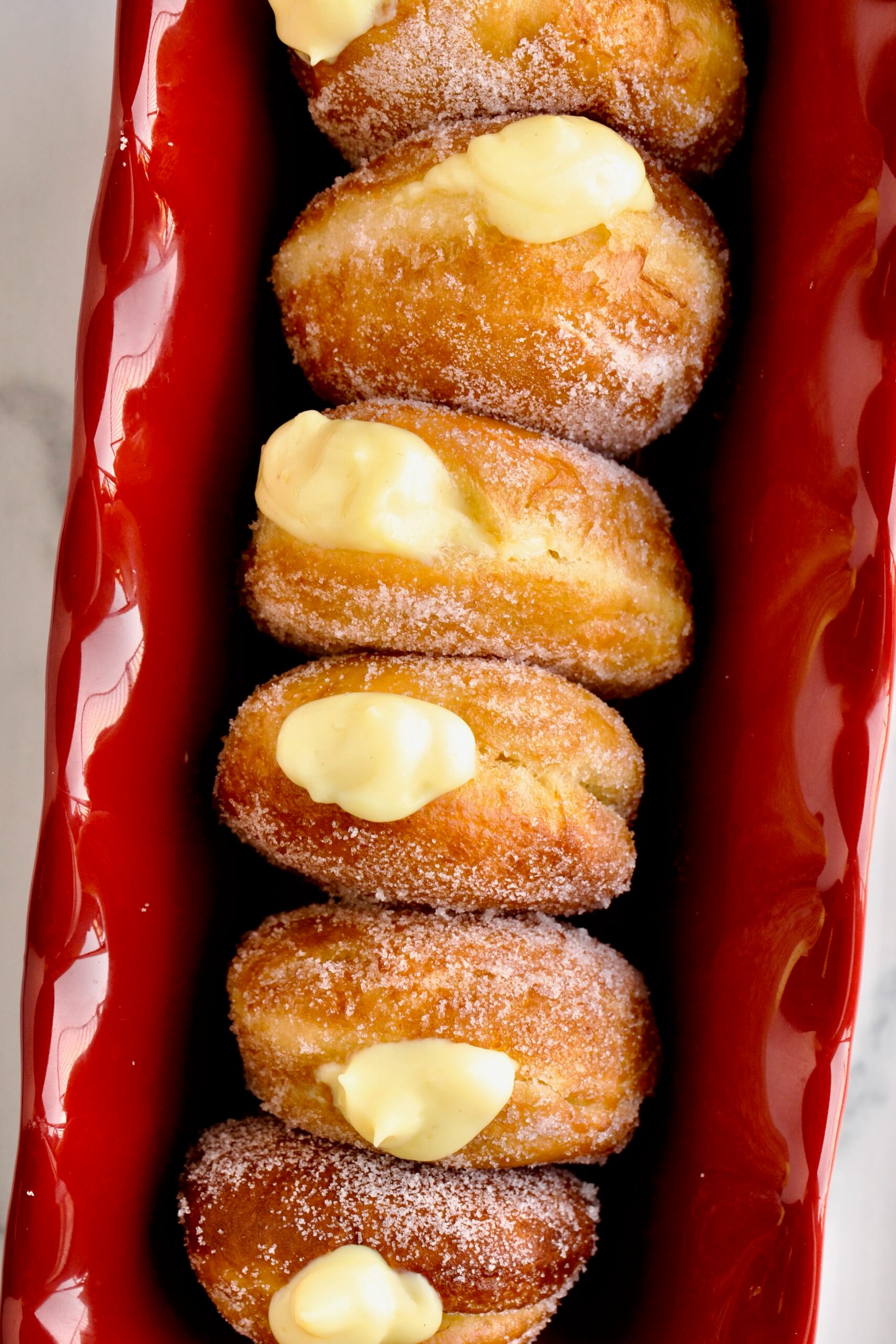 Bomboloni Recipe (Italian Donuts with Cream Filling) in a red ruffled narrow cake pan. Cream coming out of the top of the donuts.