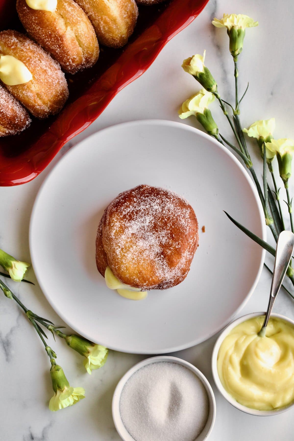 one bomboloni recipe on a plate with pastry cream in the backound and yellow flowers as decoration.