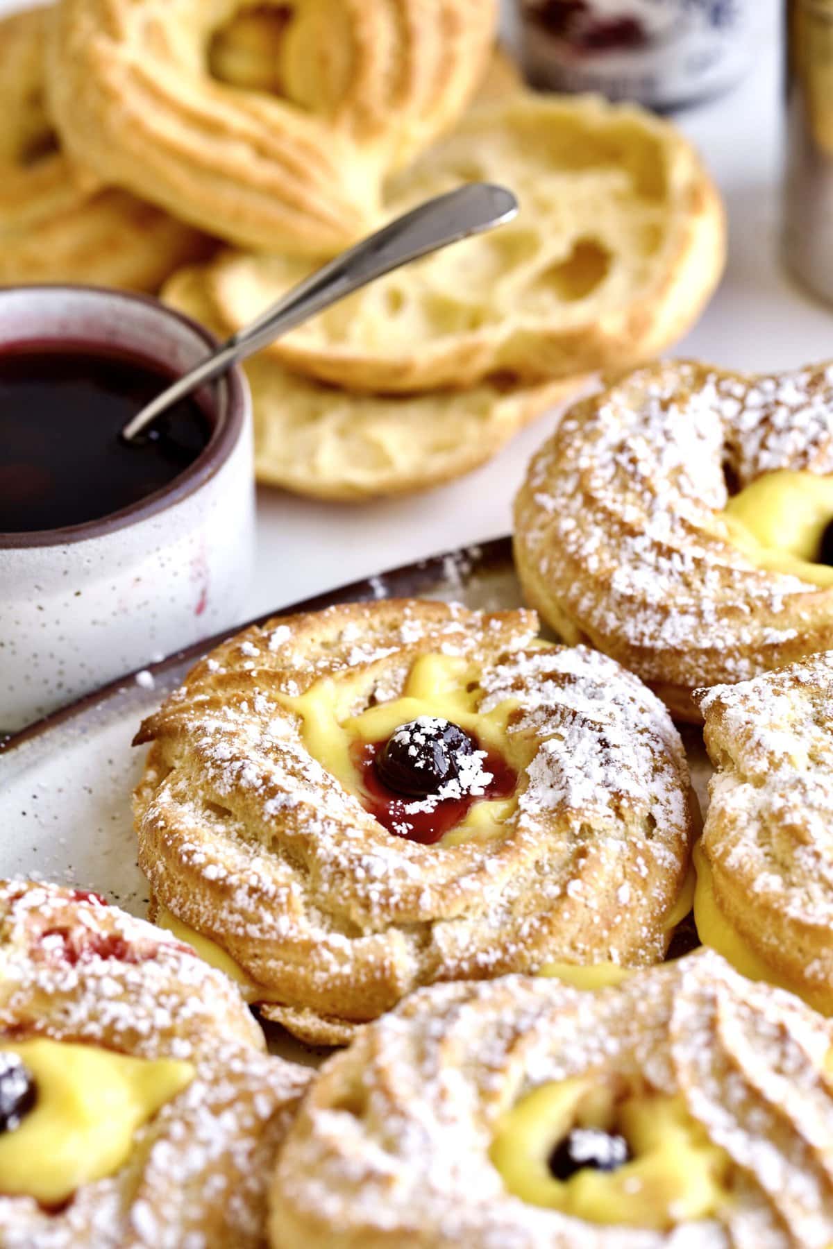 close up of finished baked zeppole filled with Italian pastry cream and Italian Amarena cherries. In the background there is a stack of unfilled zeppole and cherries.