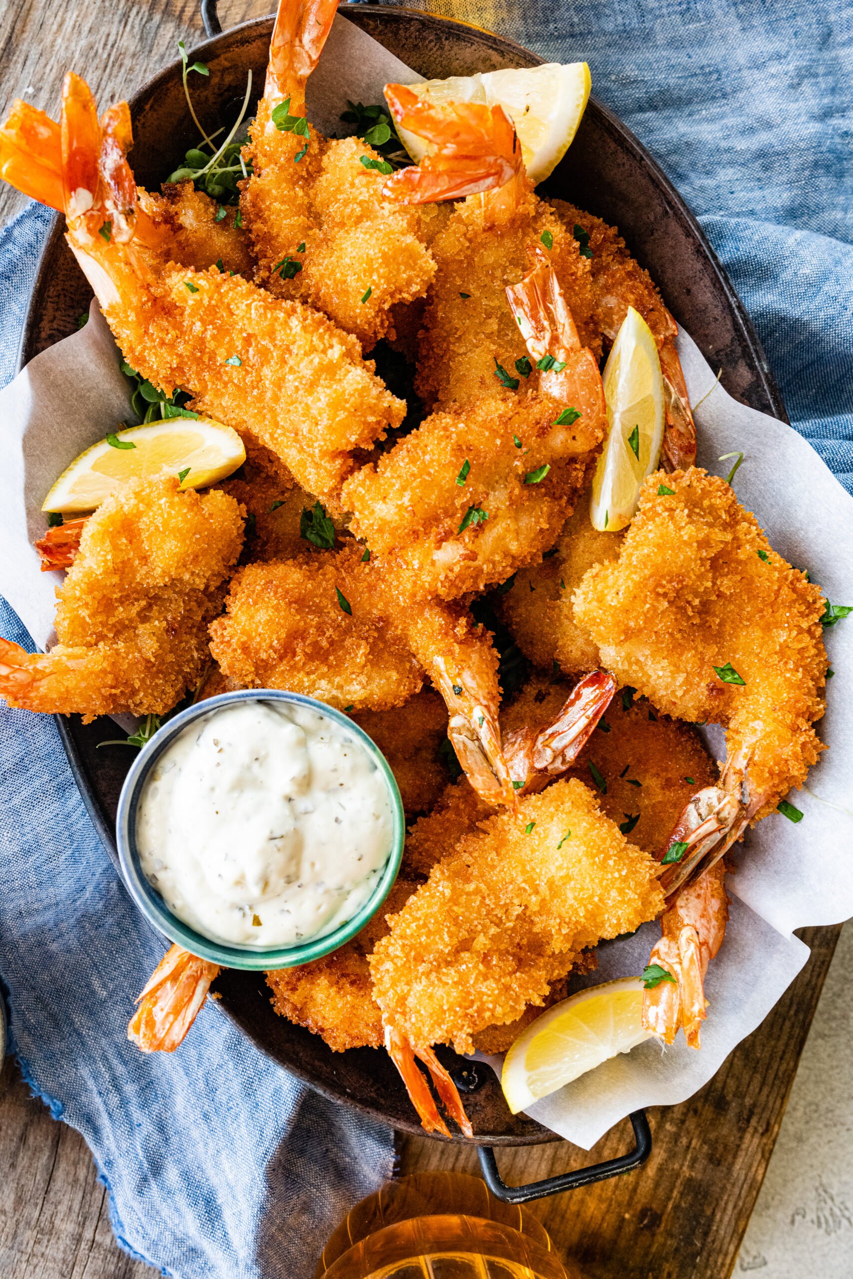 Fried shrimp on serving platter with lemon wedges, parsley, and tartar sauce. Blue napkin on the side. 