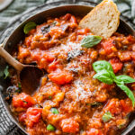 Italian Sautéed eggplant with tomatoes in a serving platter with wooden spoon and side of ciabatta bread.