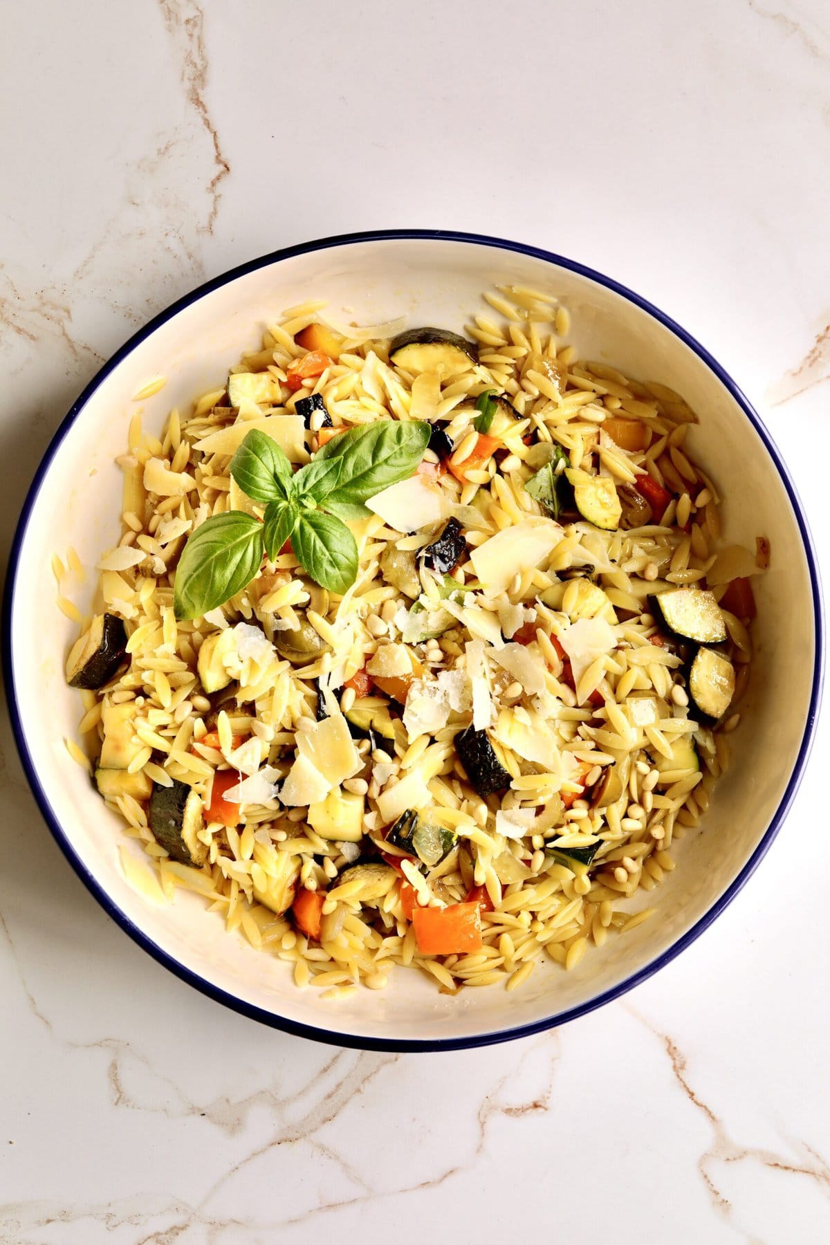 easy orzo pasta with roasted veggies in a large serving bowl. Thick slices of Parmesan cheese on top. Basil to garish.