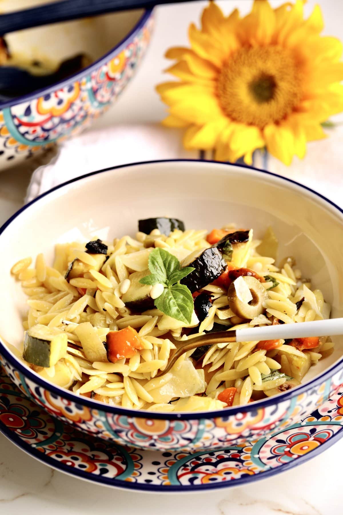 big bowl of orzo pasta with roasted vegetables with a fork ready to eat. Flowers in the background. 