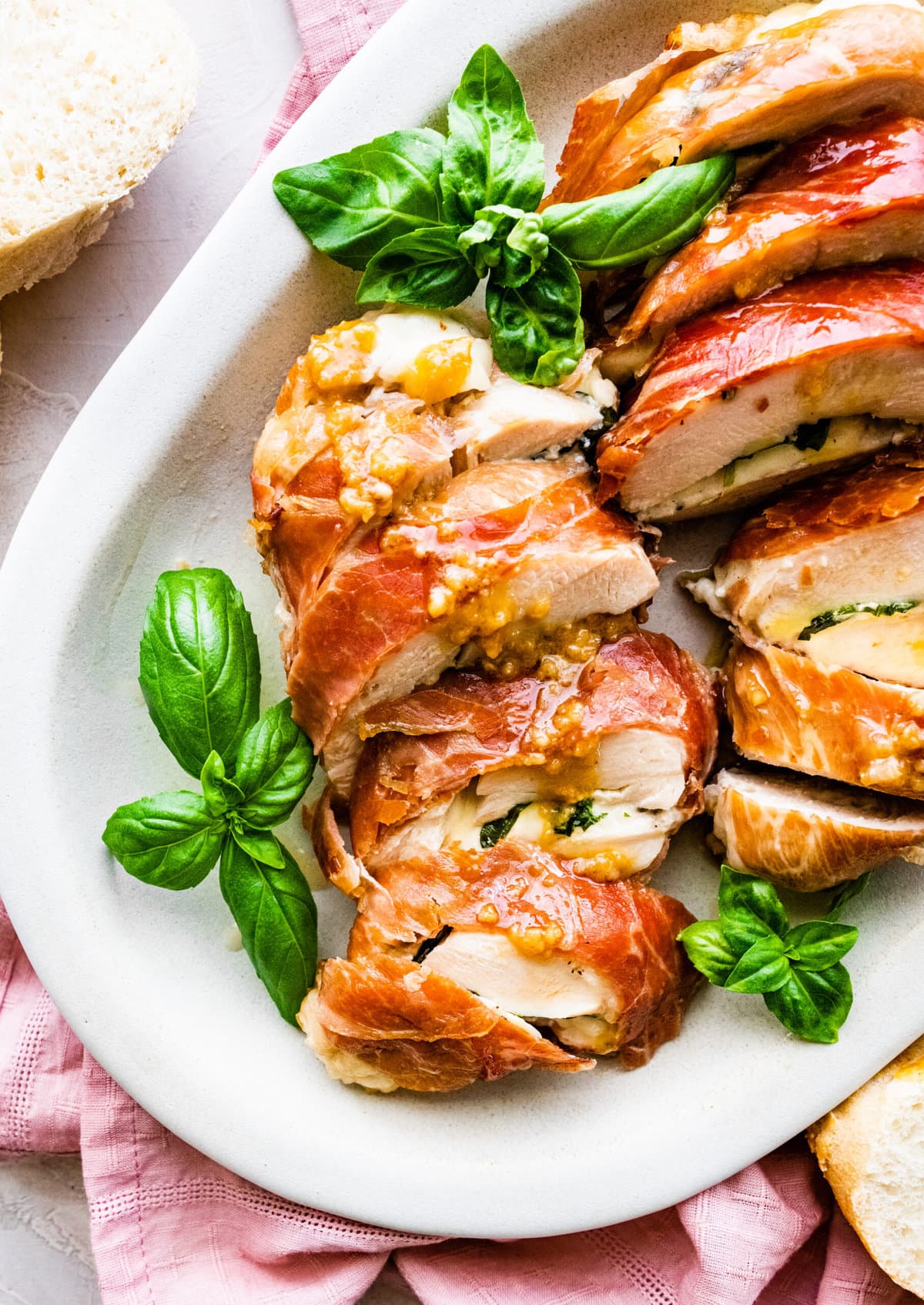 How to make Mozzarella stuffed Chicken chicken- finished stuffed chicken cut in slices and served on a white platter with fresh basil. Drizzled with pan sauce over the top. Bread slices in background.