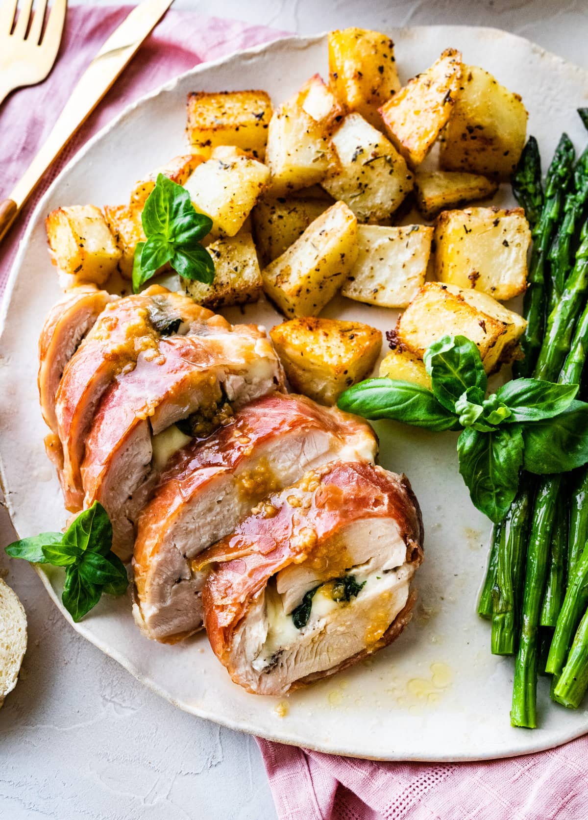 How to make Mozzarella stuffed Chicken chicken- finished stuffed chicken cut in slices and served on a white platter with fresh basil. Bread slices in background. Served with roasted potatoes and asparagus. Fork and knife on plate.