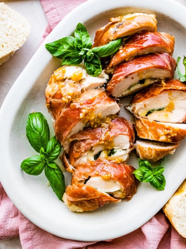 How to make Mozzarella stuffed Chicken chicken- finished stuffed chicken cut in slices and served on a white platter with fresh basil and drizzled with pan sauce. Bread slices in background. Served with roasted potatoes and asparagus.