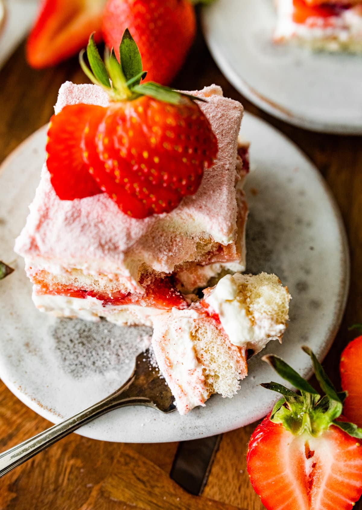 Single slice of the strawberry tiramisu on a plate with a sliced strawberry on top on a plate. A fork on a plate ready to eat. Bite taken out of one slice.