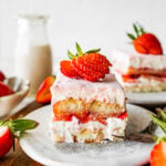 Single slice of the strawberry tiramisu on a plate with a sliced strawberry on top on a plate. Cover photo.