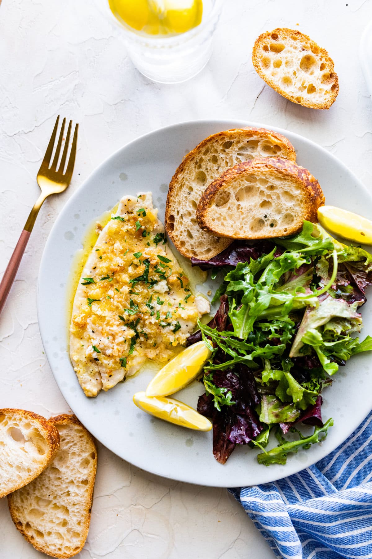 Baked flounder with butter garlic sauce on a plate with a green salad and crusty bread. 