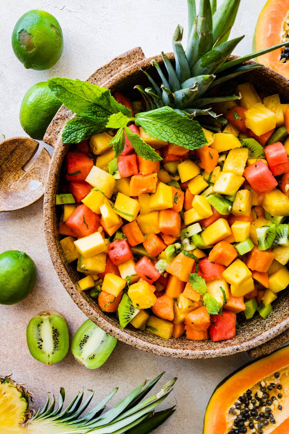 Big bowl of Best and Easy Mexican Fruit Salad in a large serving bowl with mint on top. Top of pineapple added for a festive twist. Papaya halves around the bowl. Kiwi and limes around the bowl.