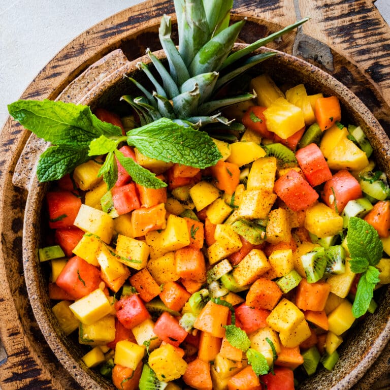 Big bowl of Best and Easy Mexican Fruit Salad in a large serving bowl with mint on top. Top of pineapple added for a festive twist. Papaya halves around the bowl.