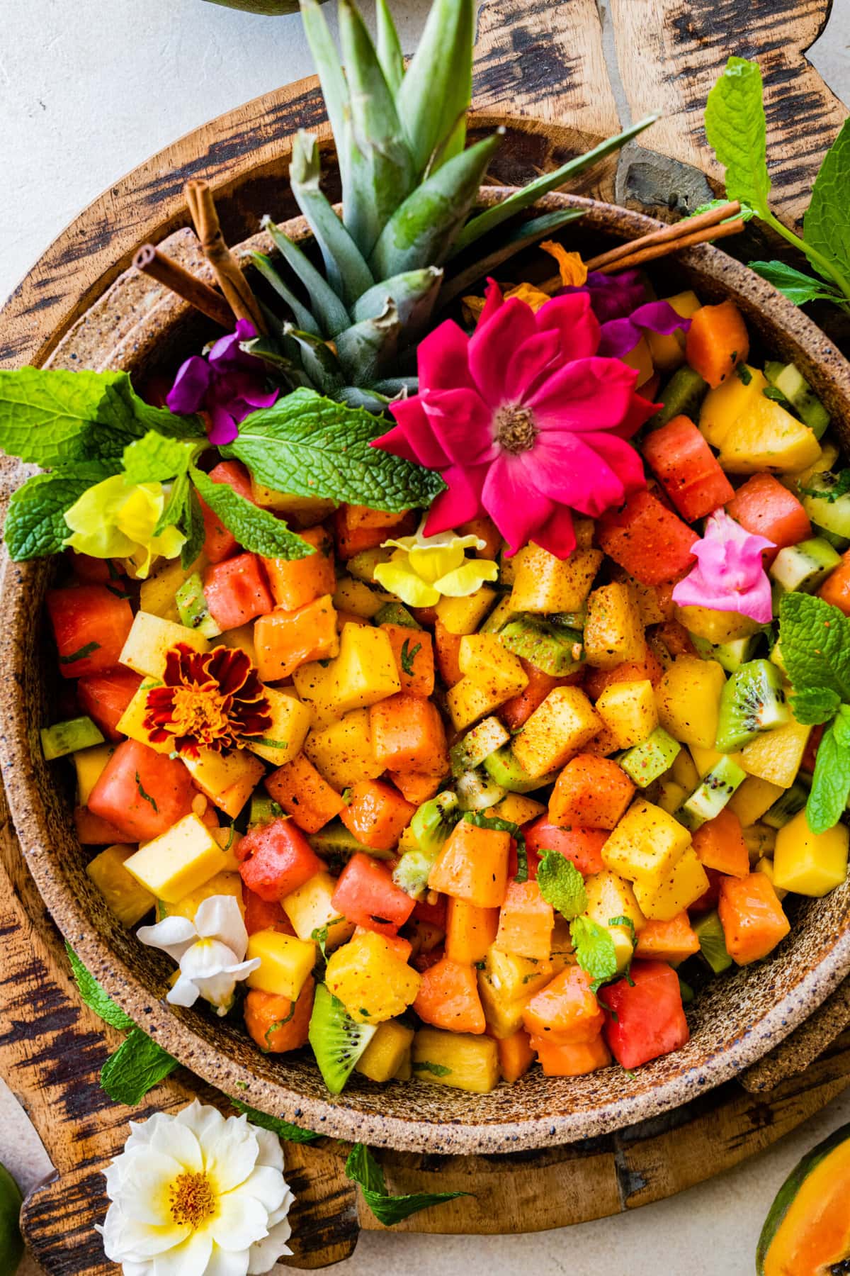 Big bowl of Best and Easy Mexican Fruit Salad in a large serving bowl with festive flowers around the bowl with mint and kiwi. Top of pineapple added for a festive twist. Papaya halves around the bowl.