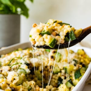 Spoon grabbing fregola sarda pasta with zucchini and cheese from a baking dish.