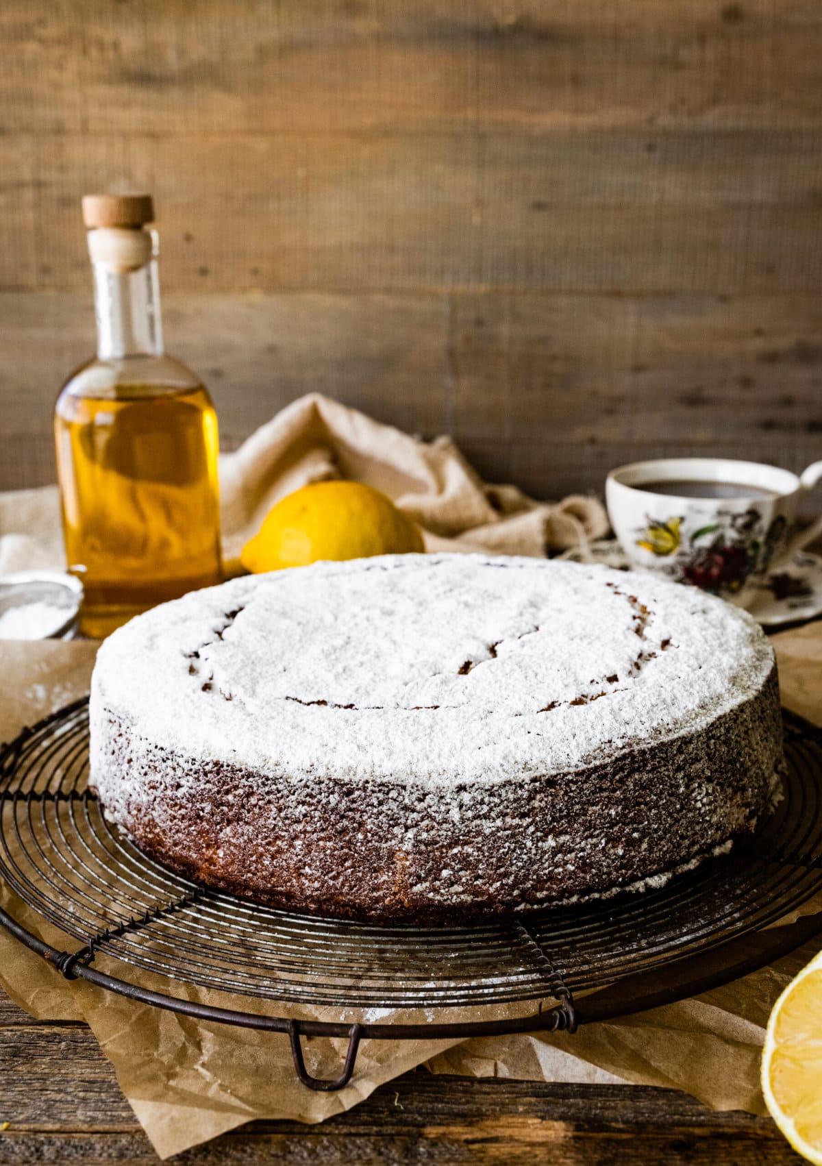 baked best olive oil cake with powdered sugar on a cooling rack with lemons around and bottle of olive oil.