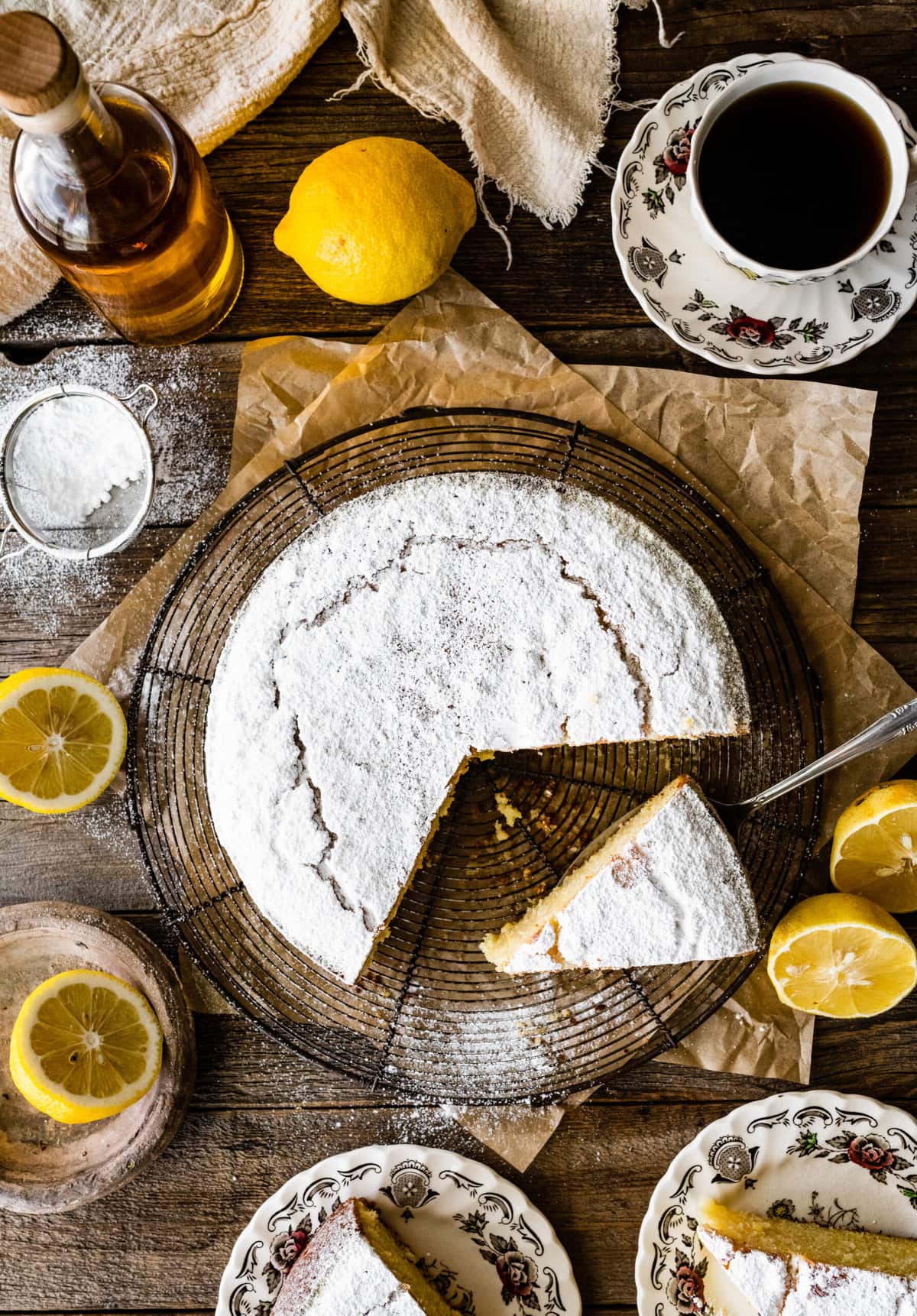 baked best olive oil cake with powdered sugar on a cooling rack with lemons around and bottle of olive oil. Slice taken out of cake.