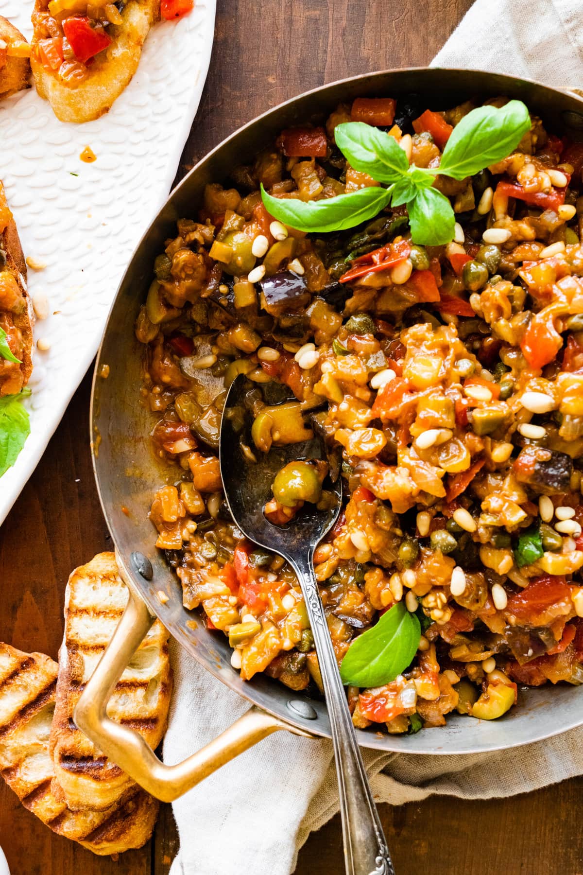 caponata in a pan with fresh basil as garnish. wood spoon in the pan.