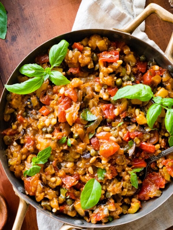 caponata in a pan with fresh basil as garnish. wood spoon in the pan.