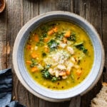 finished white bean soup in a bowl with parmigiano cheese on top on a wood background. Pieces of bread around the bowl.