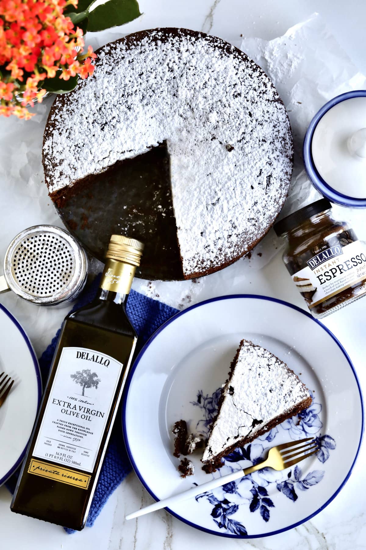 Slice of chocolate cake on a plate with blue flowers. Olive oil bottle and whole cake in background.